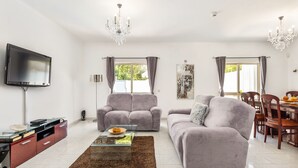 A modern living room with a flat-screen TV, two grey sofas, a glass coffee table, and a dining area with wooden chairs.