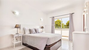 A bedroom with a bed, bedside tables, a nightstand, a dresser, a window with curtains, and a balcony.