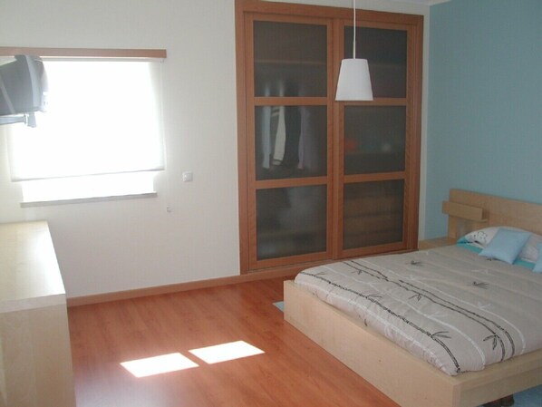 Ein Schlafzimmer mit Holzboden, einem Bett mit gemustertem Überwurf, einem Kleiderschrank mit Glastüren und einem Fenster mit Vorhängen.