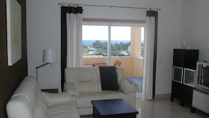 A living room with a sofa, a coffee table, a TV stand, and a view through a large window. - Luxury quiet apartment with private pool near the beach and mountains in Algarve (Portimao)