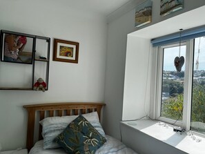 A bedroom with a bed, a window with a heart-shaped decoration, and a wall-mounted shelf with a teddy bear and a framed picture.