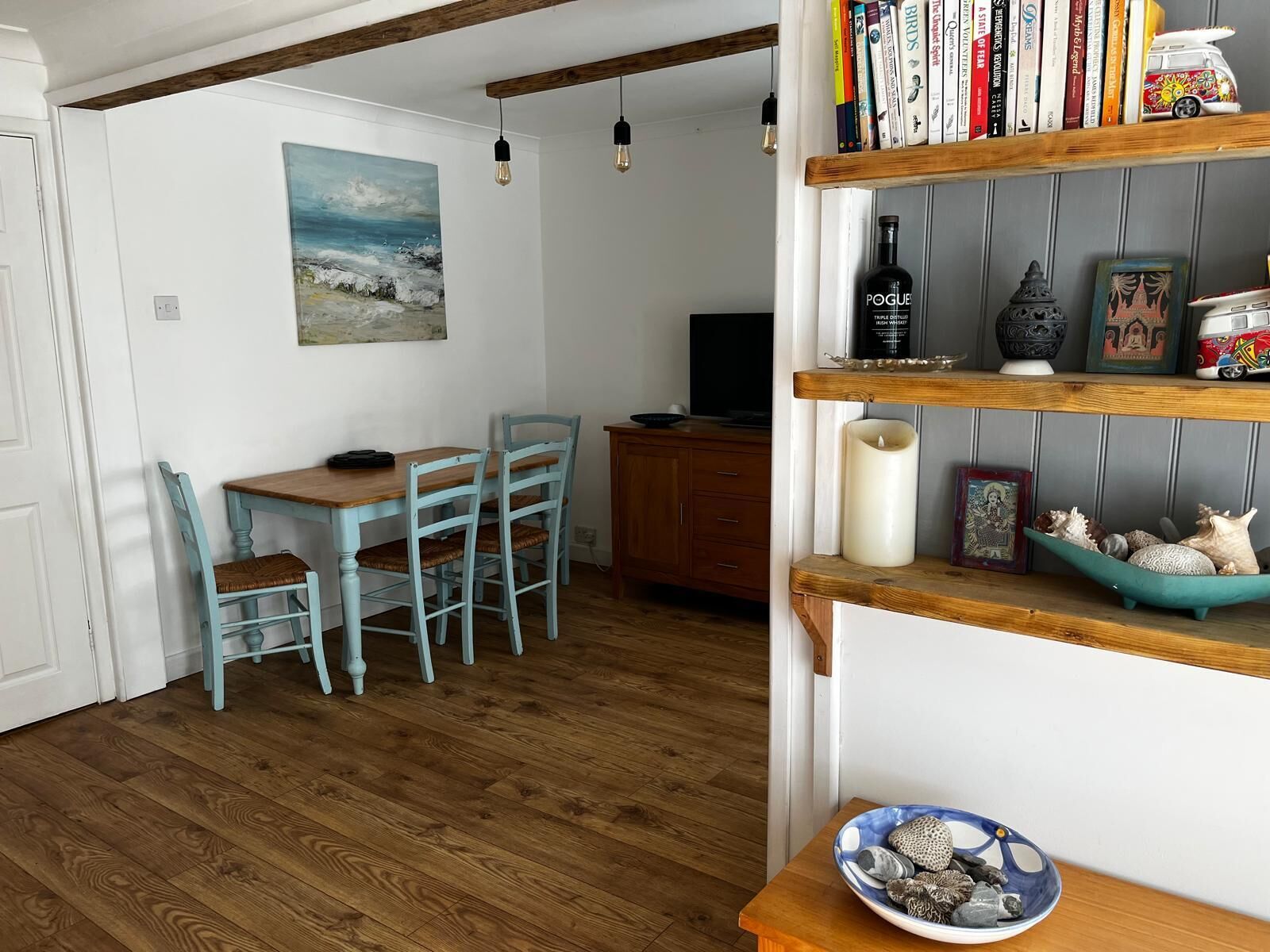A dining area with a wooden table and chairs, a painting on the wall, and a bookshelf with books and decorative items.