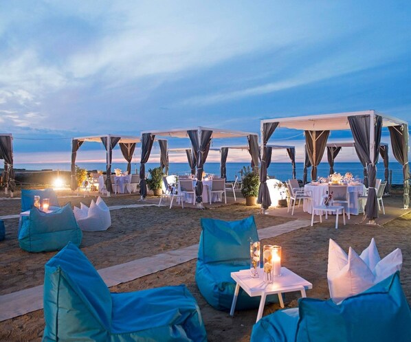 An outdoor dining area with tables and chairs, set against a beach backdrop.