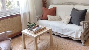 Een woonkamer met een witte bank, een klein houten tafeltje, een rond geweven vloerkleed en een raam met gordijnen.