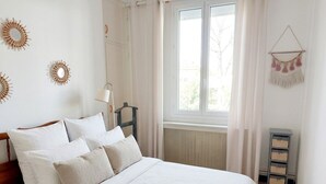 Een opgemaakt bed met witte lakens, een nachtkastje met een lamp, een raam met gordijnen en decoratie aan de muur.