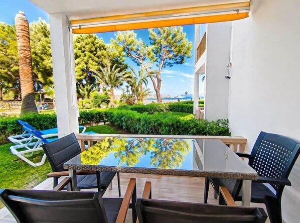 A patio with a glass table and chairs, surrounded by greenery and a view of trees and a building.