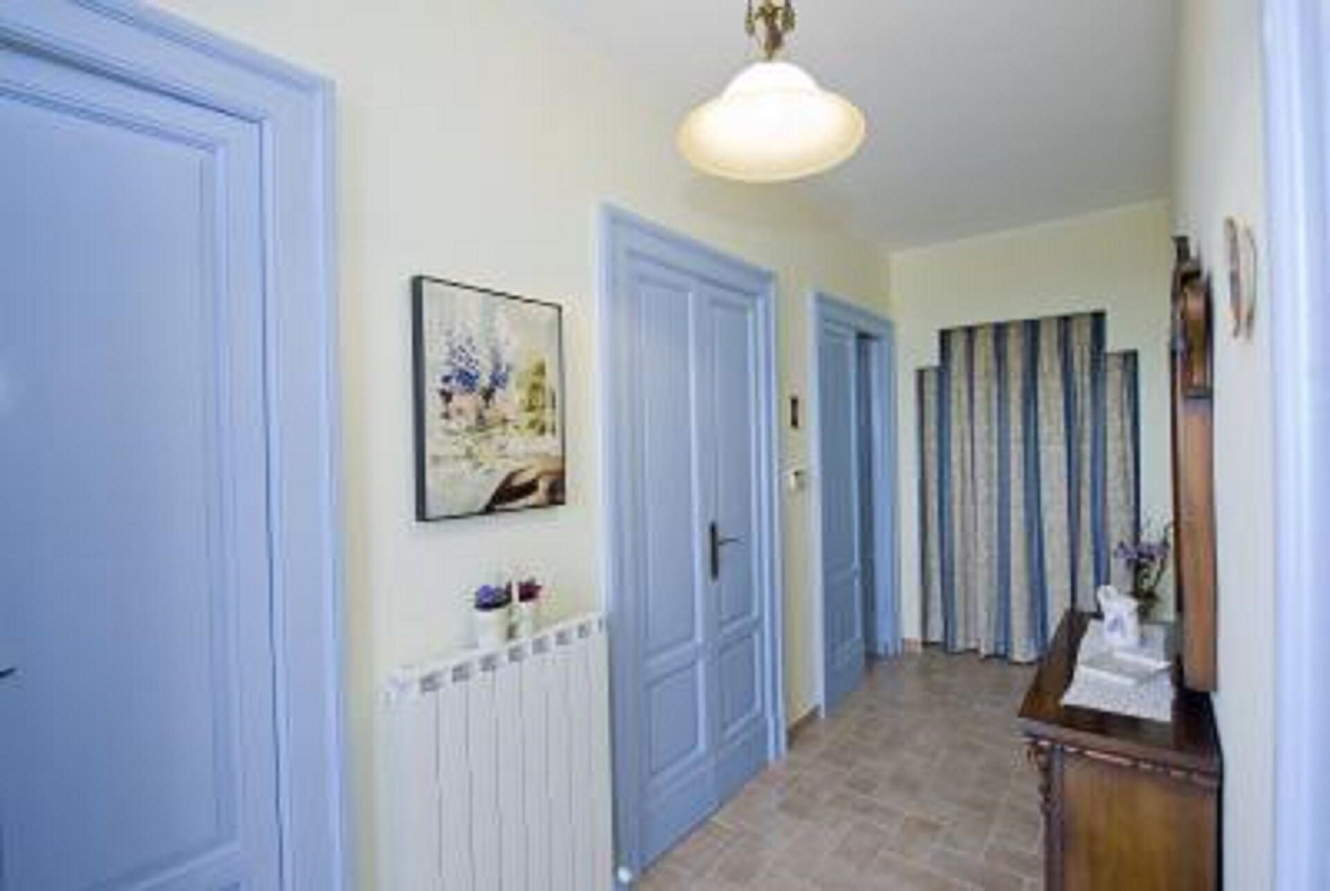 Un pasillo con puertas azules, un radiador, un cuadro enmarco y una mesa de madera con un florero.