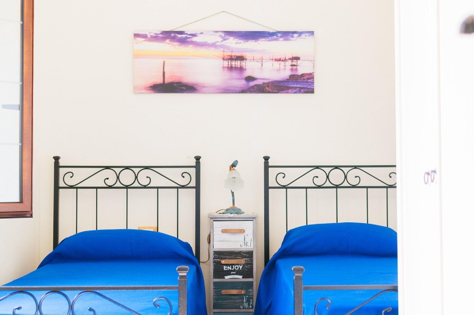 Une chambre avec deux lits, chacun recouvert d’une couvre-lit bleue et doté d’une tête de lit en métal. Un tableau représentant un coucher de soleil sur l’eau est accroché au mur, et une petite table, sur laquelle se trouvent une lampe et un objet décoratif, est également présente.