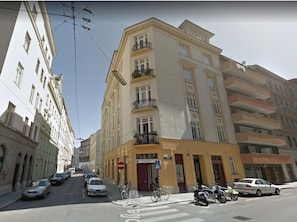 Un immeuble à plusieurs étages avec une façade jaune, un passage piéton et des voitures et des motos garées dans la rue.