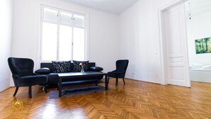 A spacious room with a wooden herringbone floor, a black sofa set, two black armchairs, a small table, and a large window with white frames.