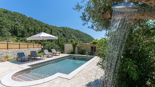 Une piscine bordée de pierres, entourée de chaises longues et d’un parasol blanc.
