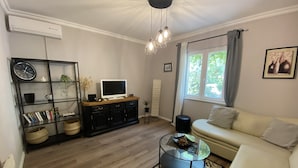 Un salon comprenant un canapé, une table basse, un téléviseur posé sur un meuble en bois, une bibliothèque, une horloge et une fenêtre avec des rideaux.