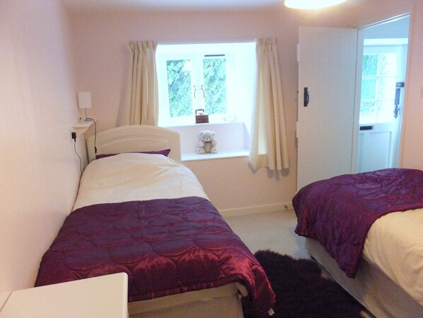 A bedroom with two beds, a window with curtains, a teddy bear on the windowsill, and a door to the right.
