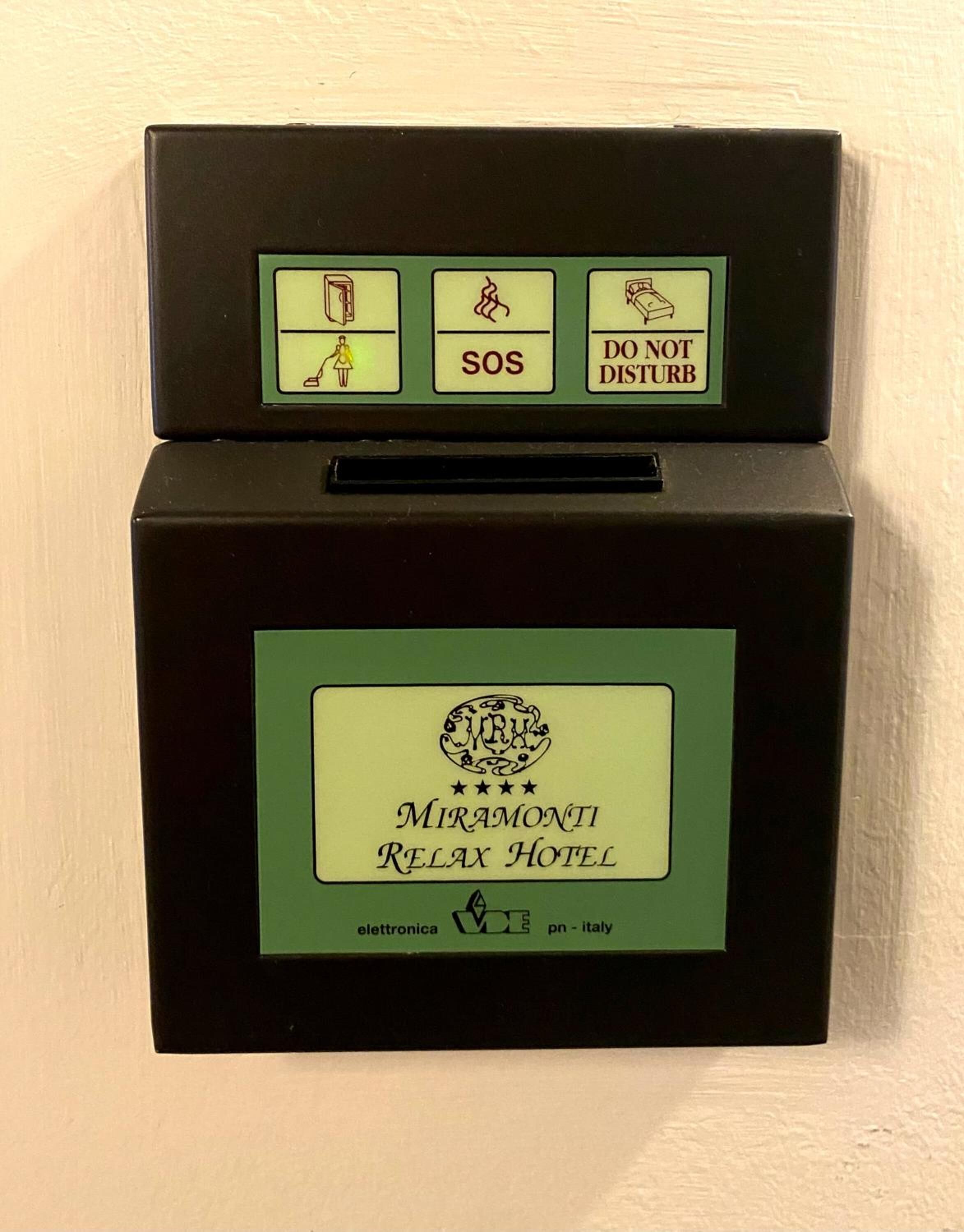 A black hotel room safe with a green label for Miramonti Relax Hotel, featuring emergency icons and a contact number.