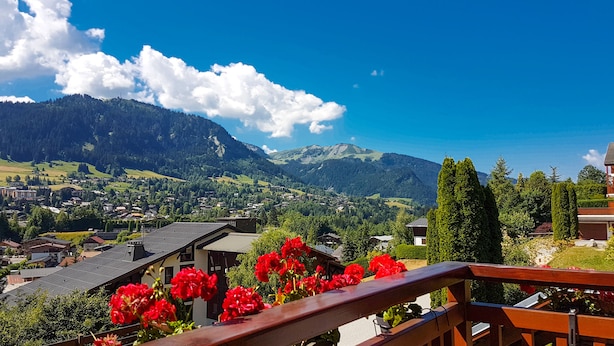 A scenic view of a mountain village with traditional houses, a wooden balcony with red flowers, and a clear blue sky.