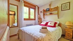A bedroom with a bed, a bedside table, a nightstand, a window with curtains, a mirror, and a shelf with decorative items.