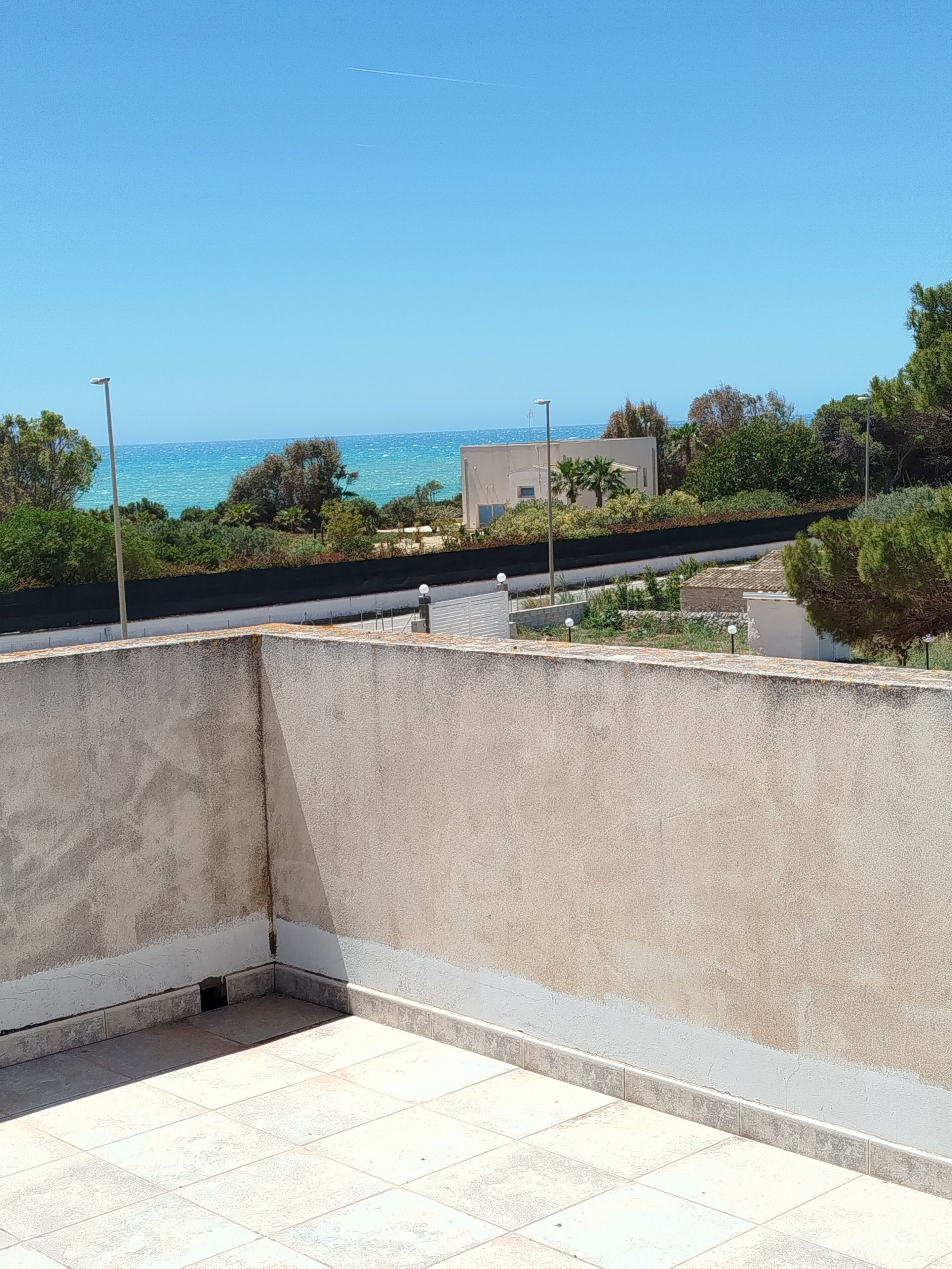 A rooftop terrace with a view of the sea, a modern building, and surrounding greenery.