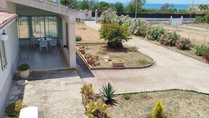 Un edificio moderno con un patio cubierto, mobiliario exterior y un jardín ajardinado con diversas plantas.