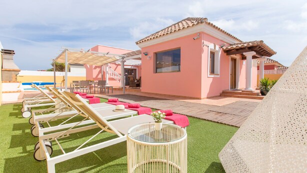 Un espace aménagé au bord de la piscine, avec des chaises longues, une petite table surmontée d’un vase, et un bâtiment à la façade rose.