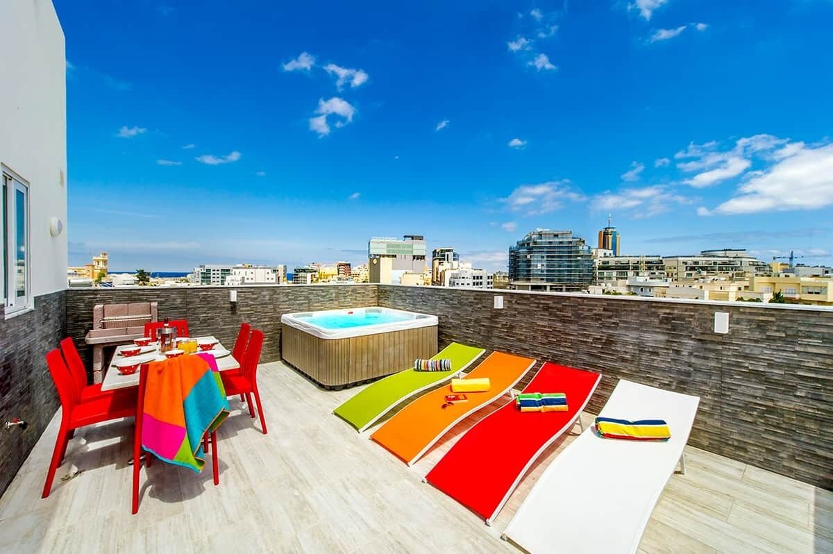 A rooftop terrace with a dining area, a hot tub, colorful mats, and a city view.