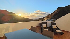 Eine Terrasse mit Tisch und zwei Loungestühlen, mit Blick auf eine Berglandschaft bei Sonnenuntergang.