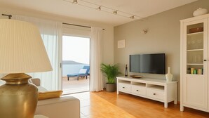 Ein modernes Wohnzimmer mit Fernseher, weißen Möbeln, einer Pflanze und Blick ins Freie.