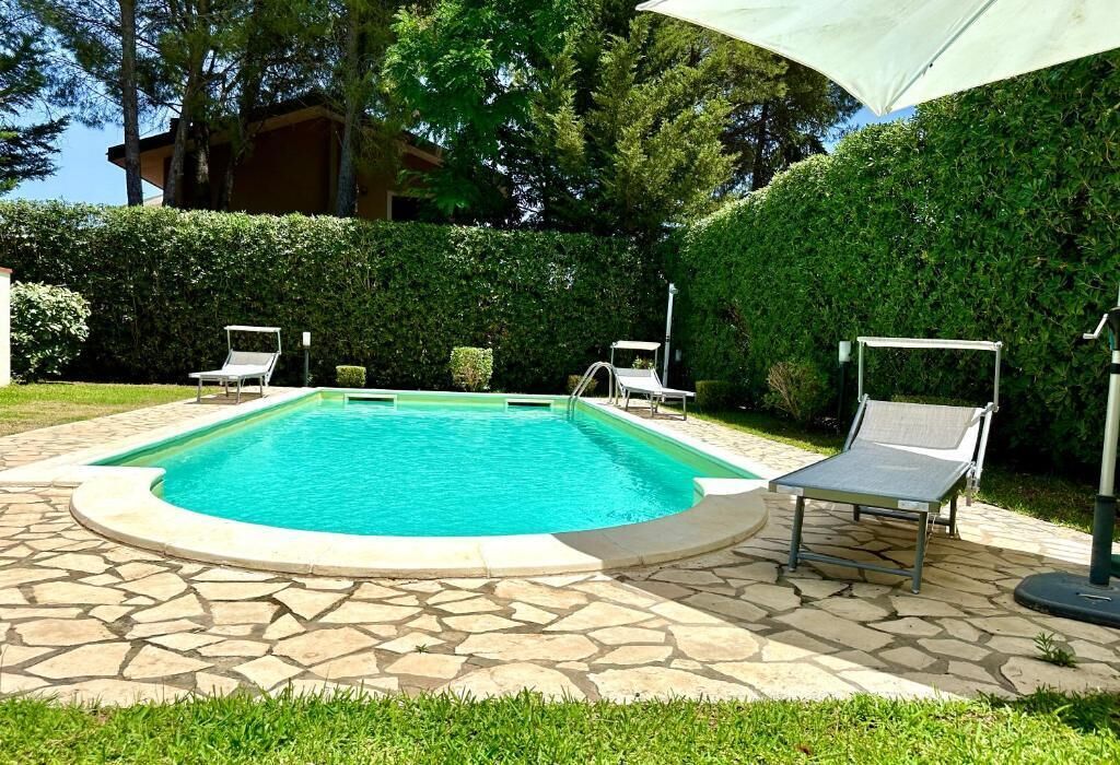 A swimming pool with a stone tile deck, surrounded by a hedge and two lounge chairs.