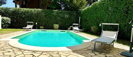 A swimming pool with a stone tile deck, surrounded by a hedge and two lounge chairs.