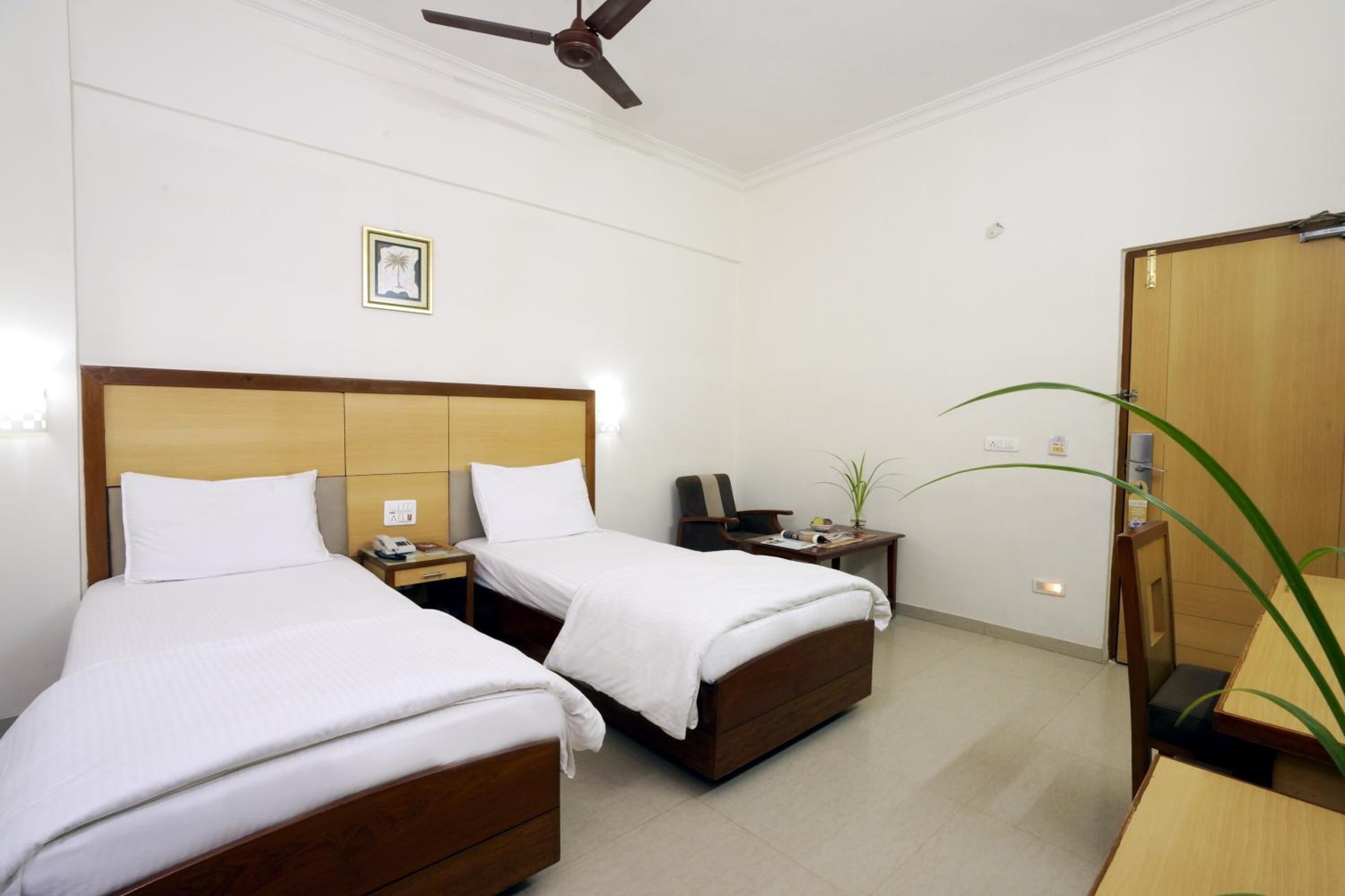 A hotel room with two beds, a desk, a chair, a small table, a ceiling fan, and a mirror.
