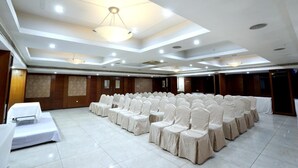 Une salle de conférence avec des rangées de chaises recouvertes de nappes blanches, une scène avec une table blanche et un éclairage encastré au plafond.