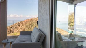 A modern living room with a large window overlooking a rocky coastline and a pool area. - Perched on a Hill Overlooking the Cretan Sea (Rethymno)