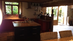 Una cocina con una isla de madera, un comedor con sillas de mimbre y ventanas amplias con vistas a zonas verdes.