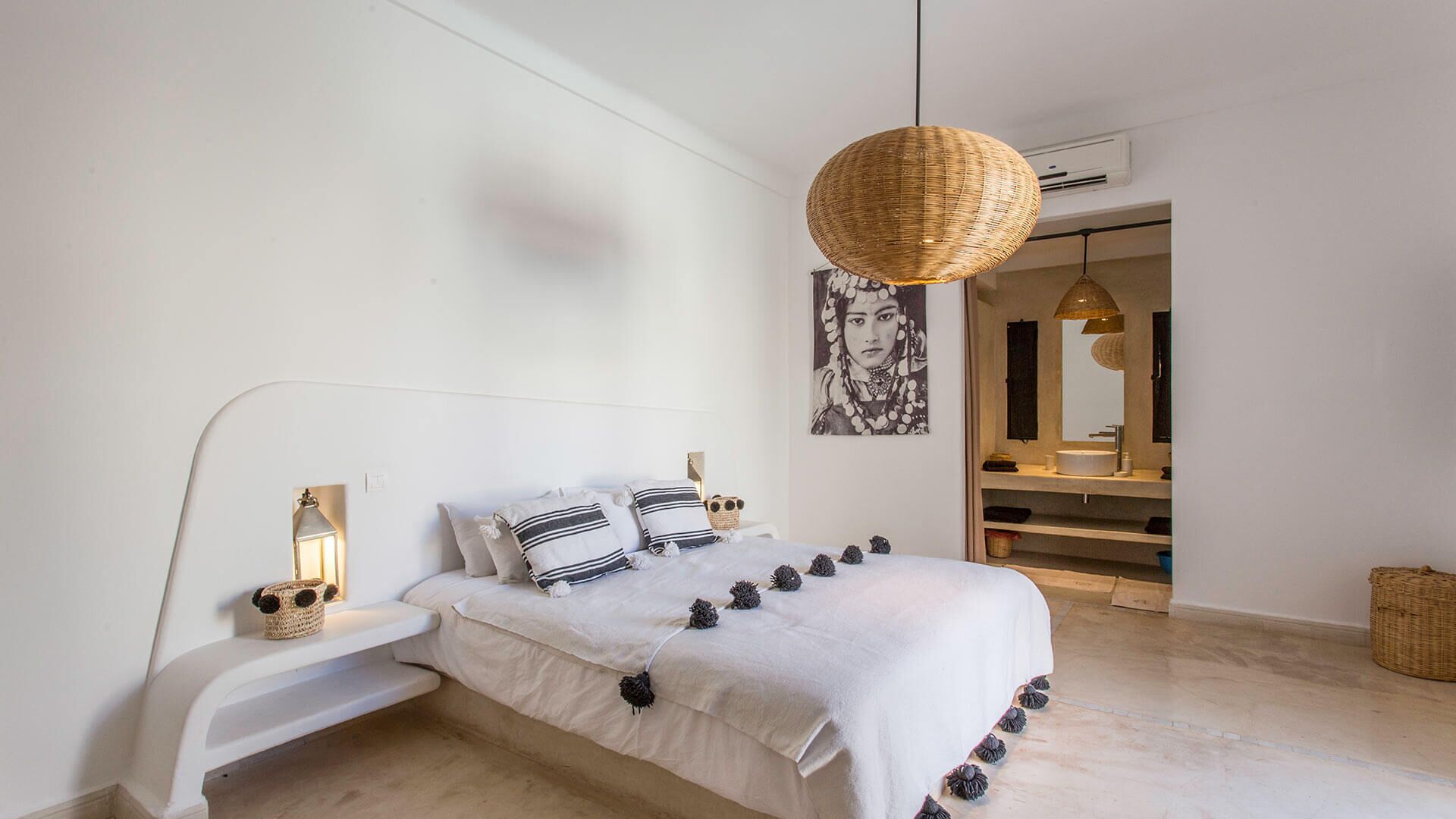 Ein modernes Schlafzimmer mit einem weißen Bett, gestreifter Bettwäsche, einer hängenden gewobenen Leuchte und einem großen Spiegel über dem Schminktisch.