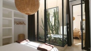 Une chambre avec un grand lit, une suspension en osier, une porte en verre donnant sur un balcon agrémenté de plantes et une vue sur l’extérieur.