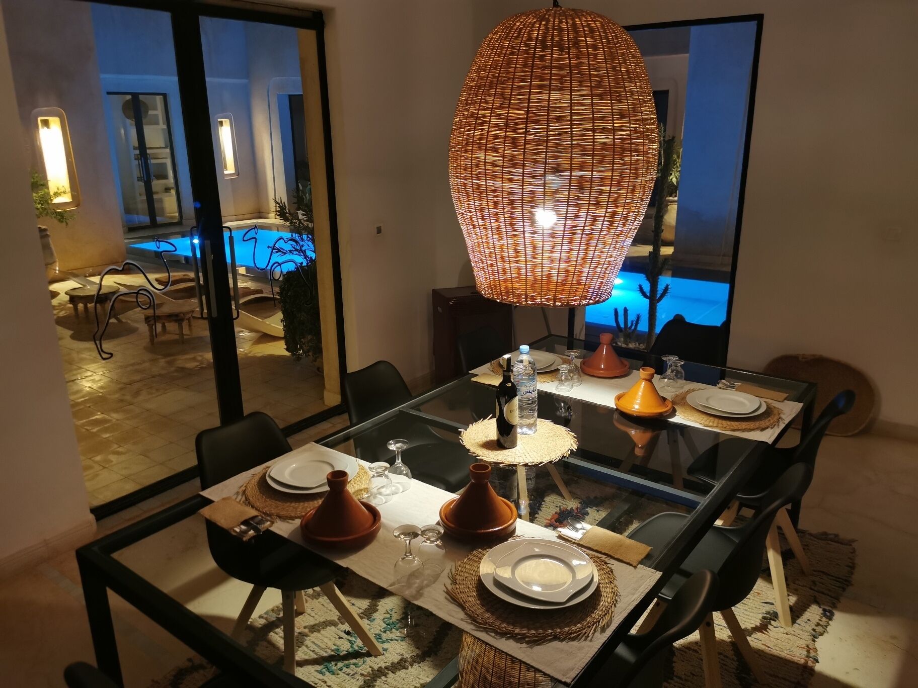 Un coin repas avec une table en verre, des chaises noires, un luminaire suspendu en fibres tressées et de la vaisselle de style marocain.