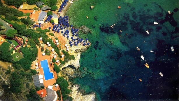 Vista aérea de un complejo turístico costero con piscina, sombrillas de playa y barcos en el agua.