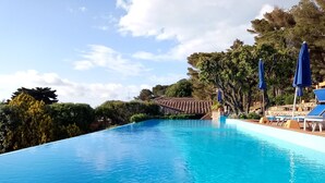 Una piscina azul cristalina con sillas de descanso y sombrillas, rodeada de árboles y un puente de piedra.