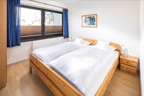 A bedroom with a wooden bed, a nightstand, a window with blue curtains, and a painting on the wall. - Birkenhof (Norderney)