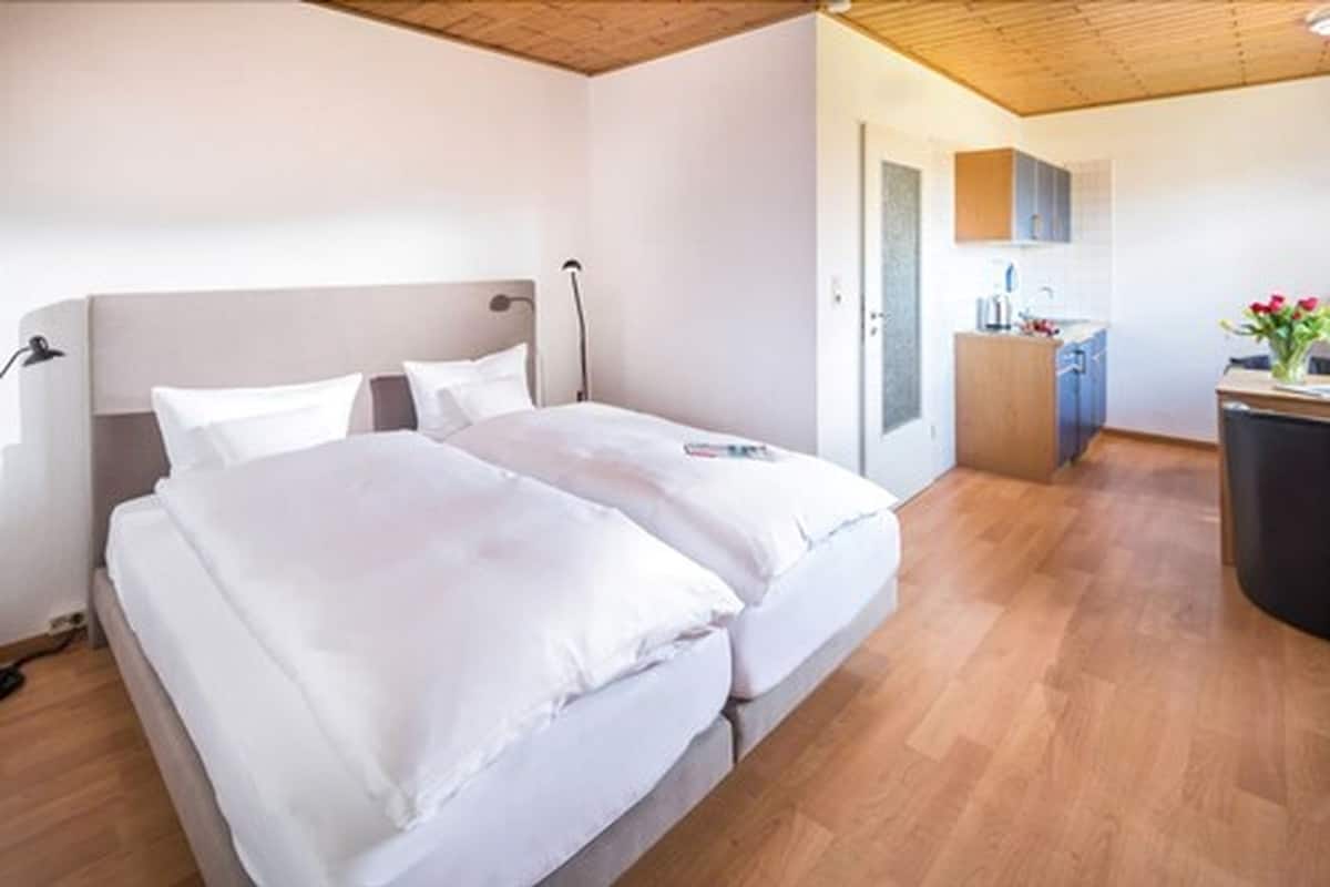 A bedroom with a double bed, a bedside table, a lamp, and a view of a bathroom and kitchen area.