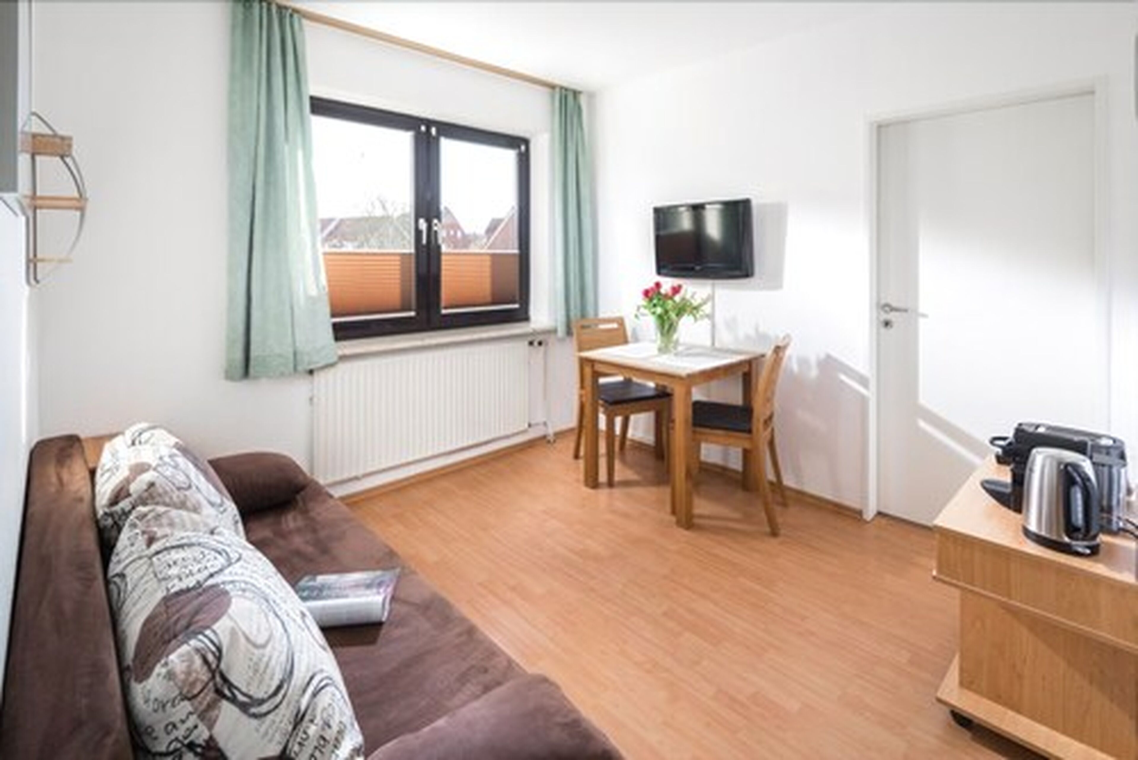 Een compacte kamer met een bruine bank, een eettafeltje met twee stoelen, een tv aan de muur en een radiator onder een raam met gordijnen.