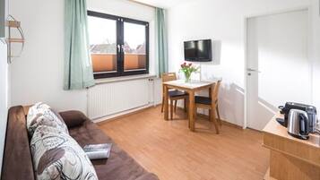 A compact room with a brown sofa, a small dining table with two chairs, a TV mounted on the wall, and a radiator under a window with curtains.