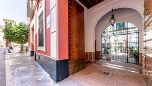 Une entrée d’immeuble avec une grande porte en bois, un sol carrelé et une fenêtre en arc surmontée d’une grille métallique.