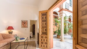 Un salon comprenant un canapé, une table basse et une lampe. Une porte en bois donne accès à un espace extérieur agrémenté de plantes et doté d’un sol carrelé.