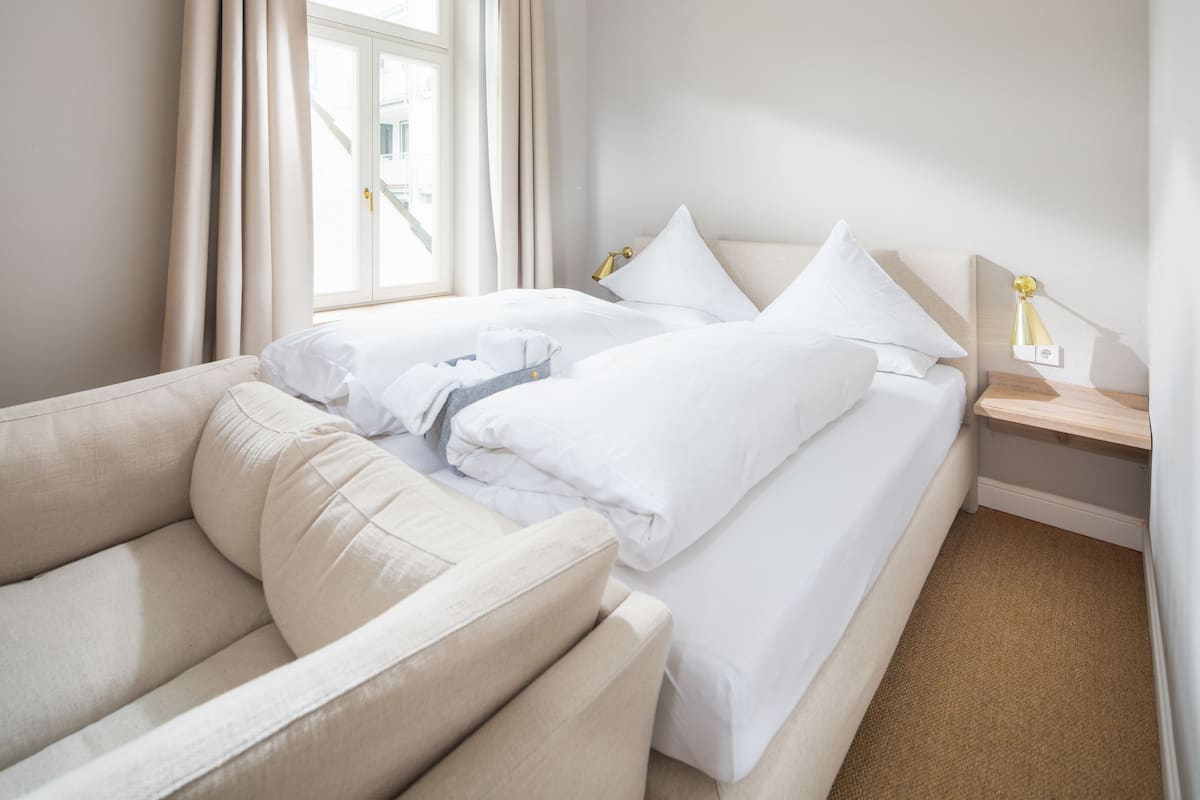 A hotel room with a bed, a sofa, a bedside table with a lamp, and a window with curtains.