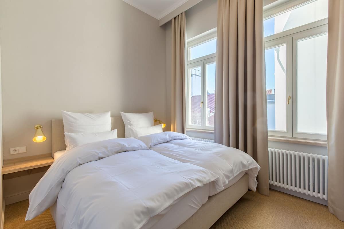 A hotel room with a large bed, two bedside lamps, a window with curtains, and a radiator.
