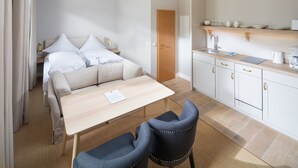 A modern hotel room with a bed, a small dining table, two chairs, a kitchenette, and a door.