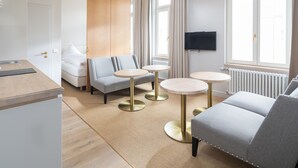 A modern hotel room with a sofa, two round tables, a TV, and a window with curtains.
