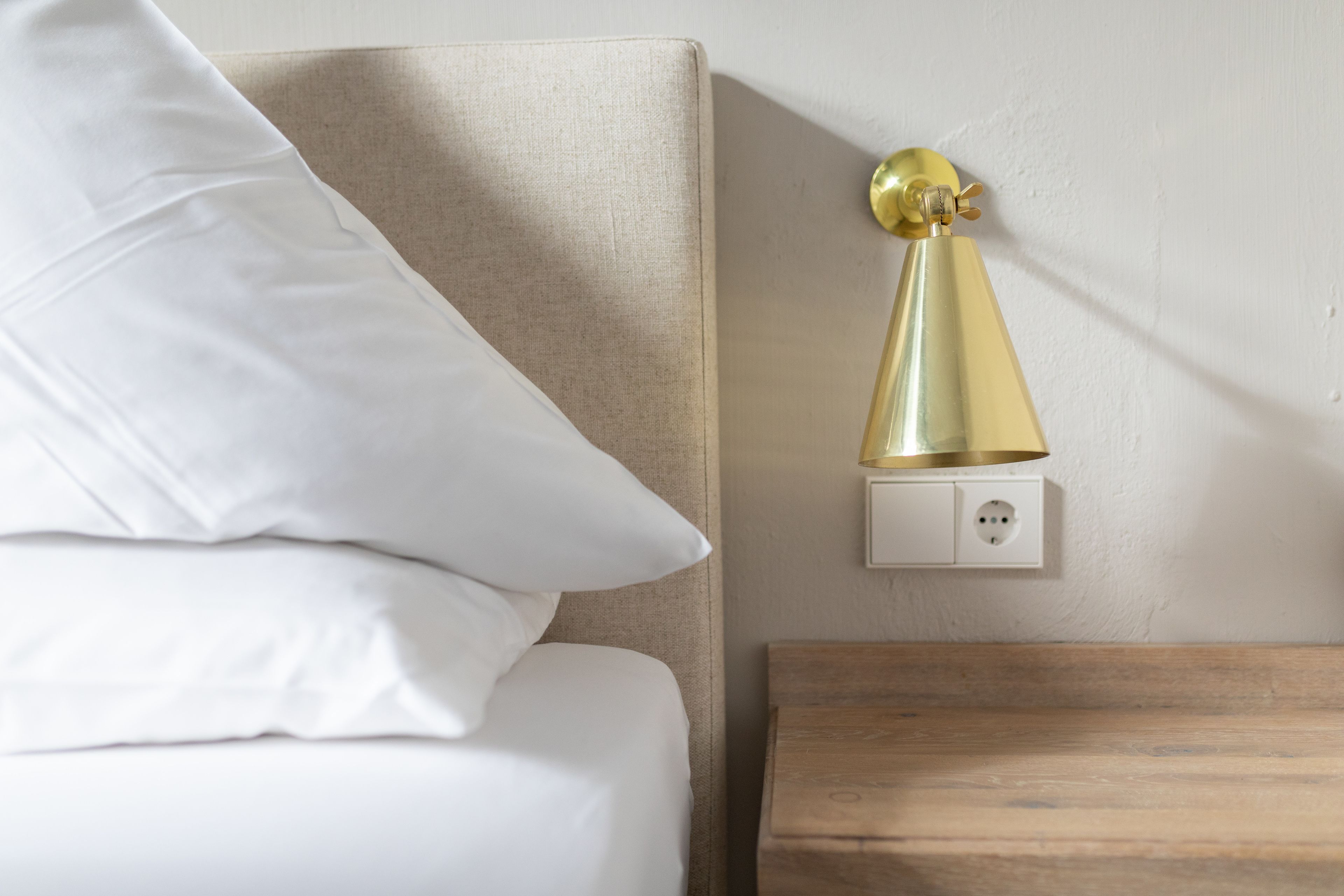 Een netjes opgemaakt bed met witte lakens en één kussen, een nachtkastje met houten afwerking en een wandlamp.
