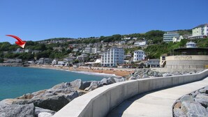 Una passeggiata costiera con marciapiede in cemento, costa rocciosa e edifici sullo sfondo.