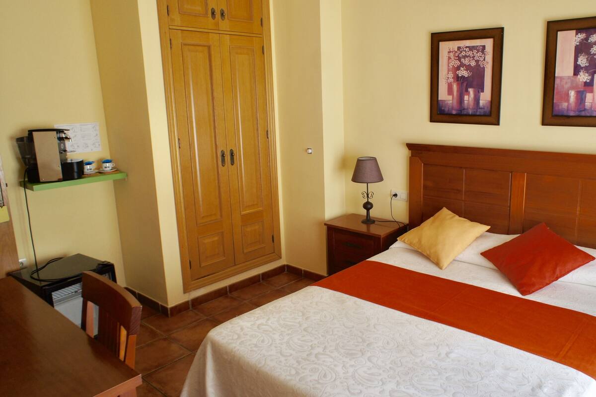 A hotel room with a wooden wardrobe, a bed with white and orange bedding, two bedside tables with lamps, and two framed floral paintings on the wall.